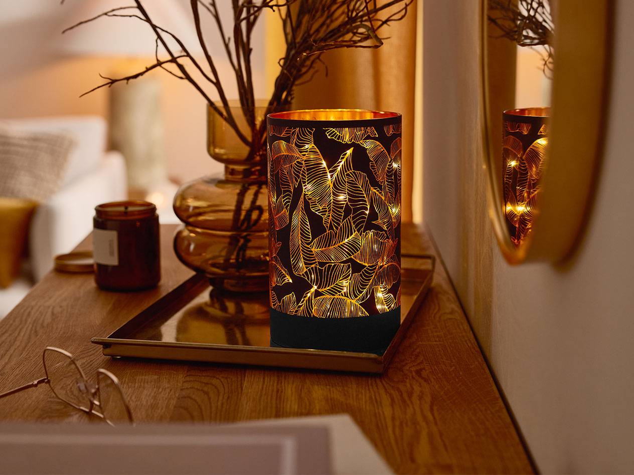 Uma lanterna LED decorativa com padrão de folhas, um vaso com galhos e uma vela em uma mesa de madeira.