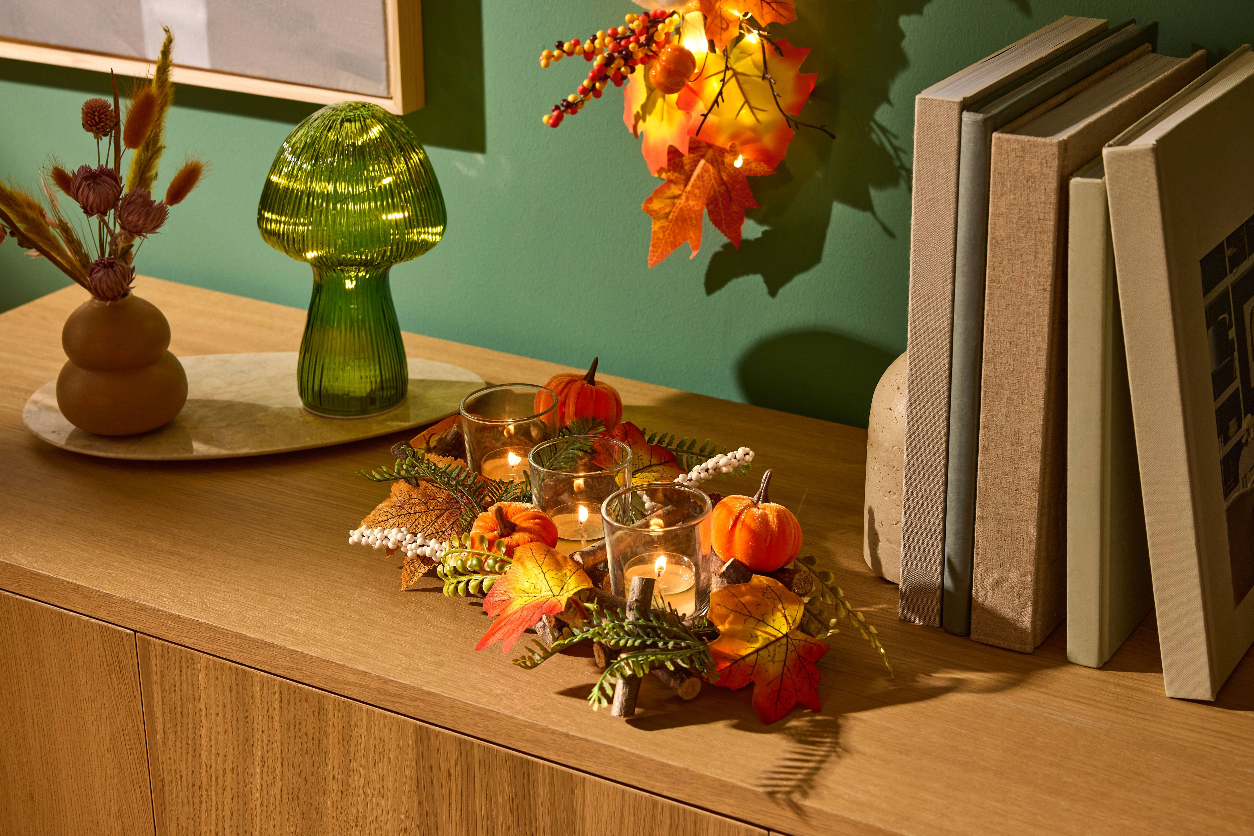 Decoração de mesa outonal com abóboras, velas e folhas, ao lado de um candeeiro cogumelo verde e livros.