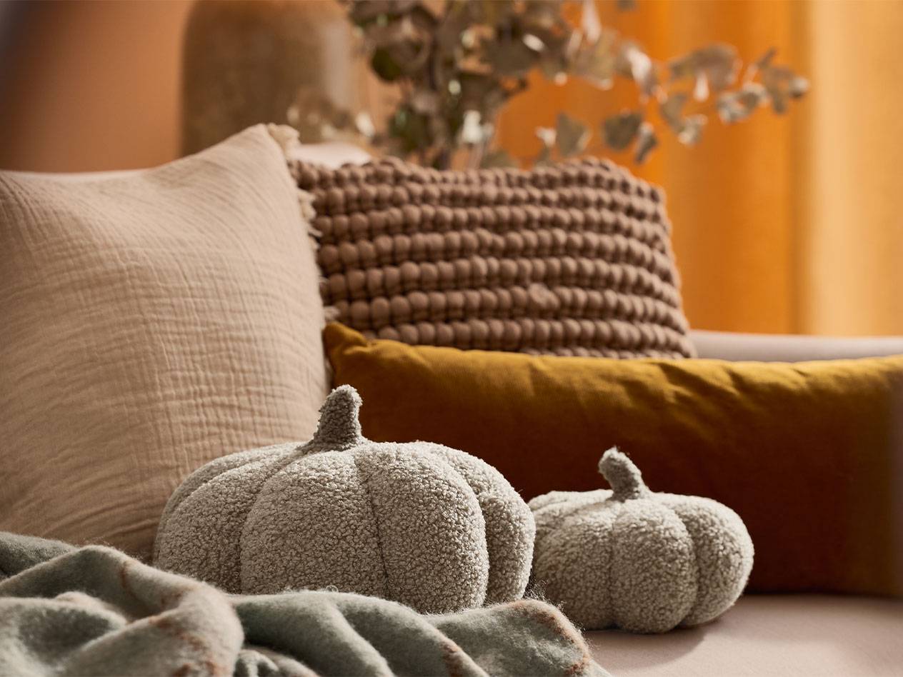 Duas abóboras decorativas de pelúcia e almofadas em tons terrosos em um sofá.