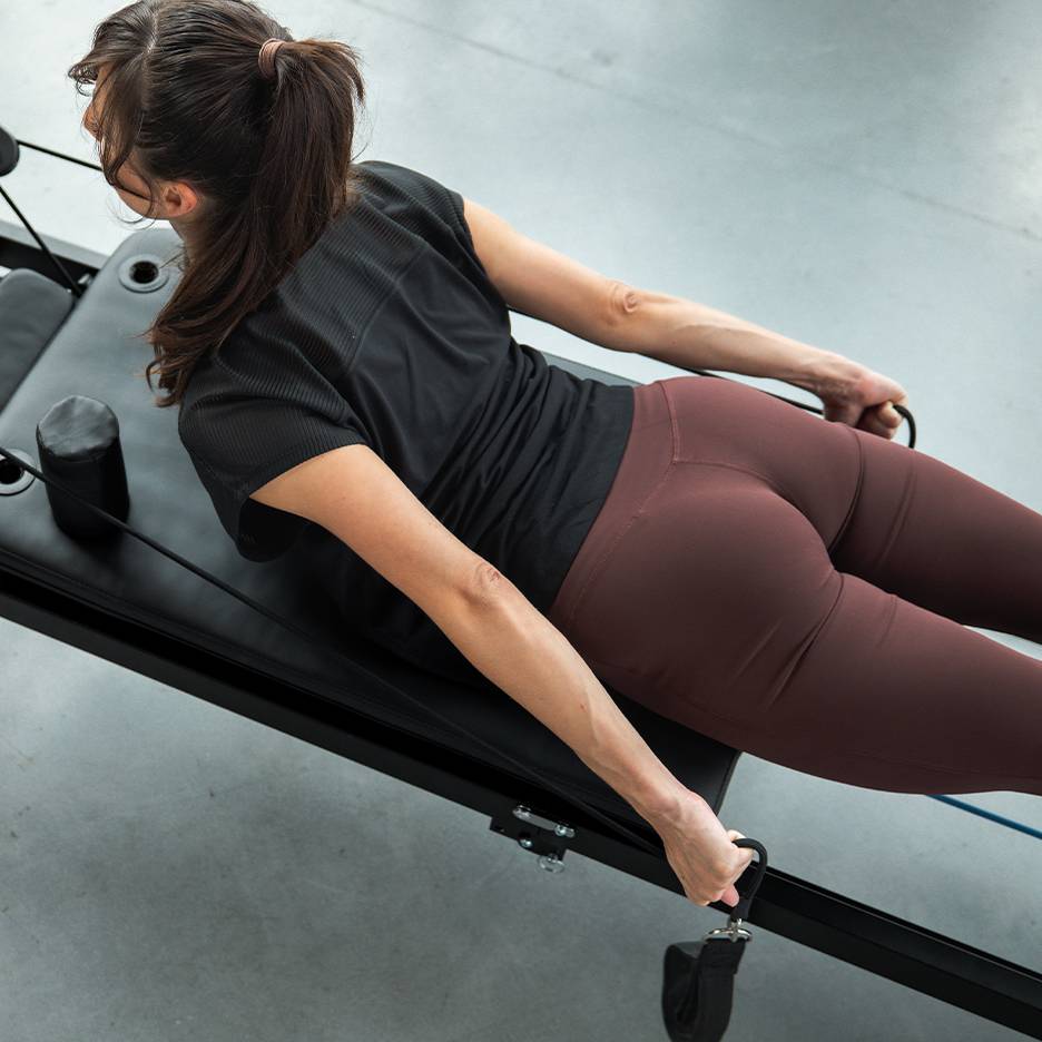 Mulher a exercitar-se numa máquina de Pilates com bandas de resistência, vestindo roupa desportiva.