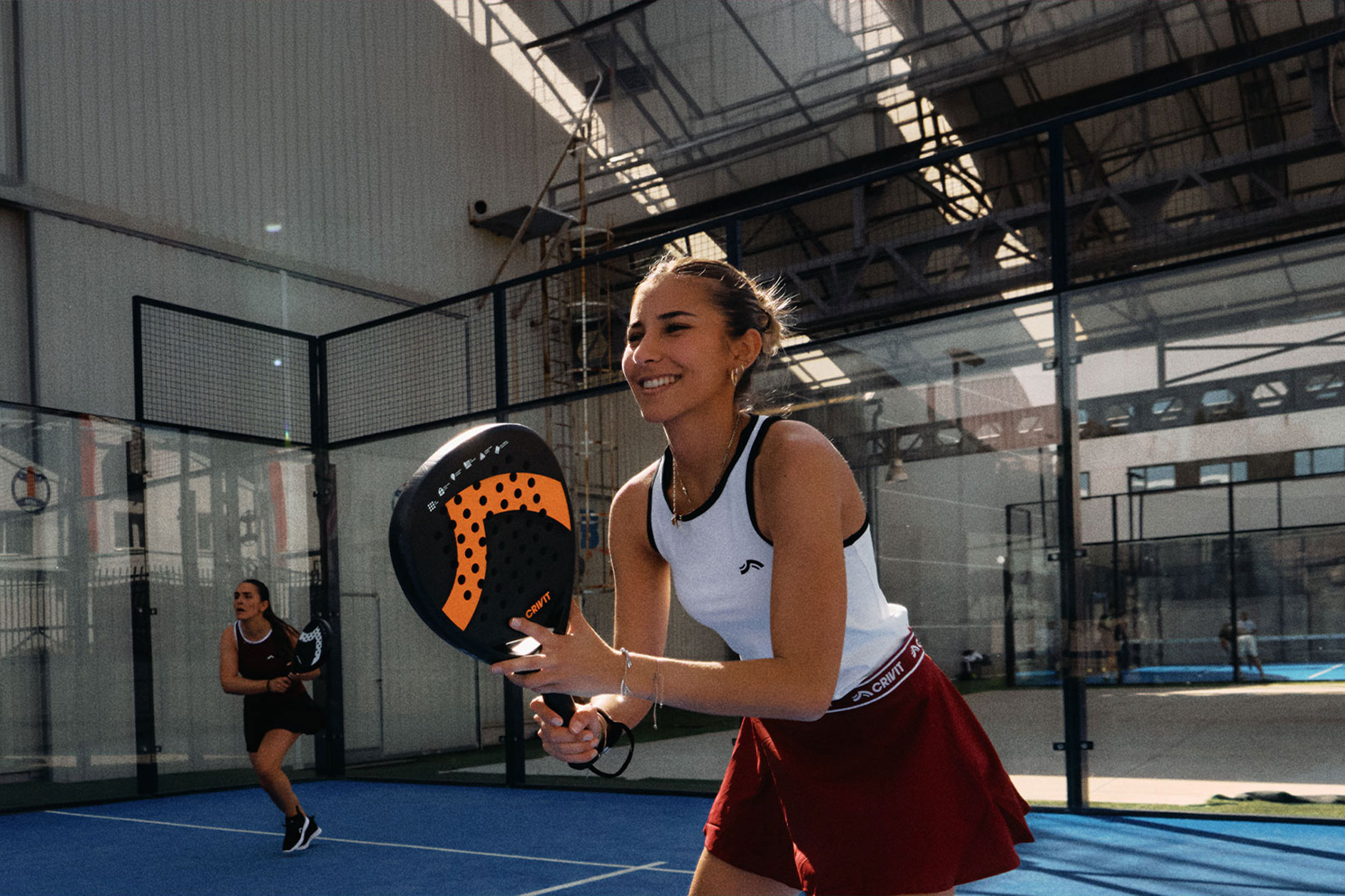 aiLabeling.aiLabelForImageAltText: Duas mulheres a jogar padel, uma a sorrir e a segurar uma raquete, num campo azul.