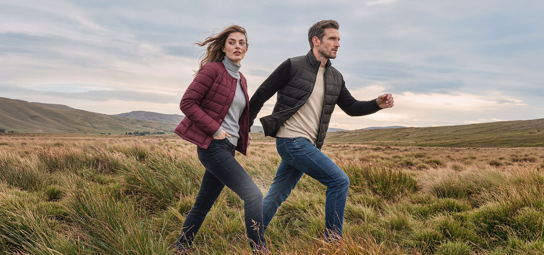 aiLabeling.aiLabelForImageAltText: Casal em casacos leves e jeans caminha por uma paisagem montanhosa e relvada.