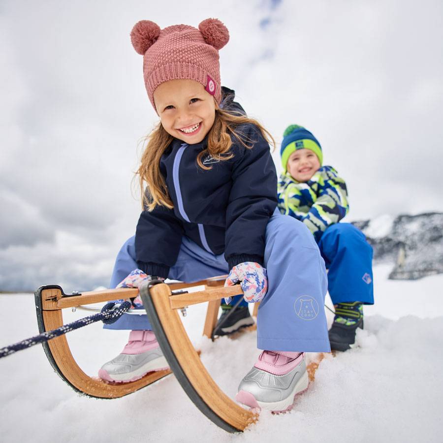 Crianças com roupas de inverno e gorros com pompons, a andar de trenó na neve.