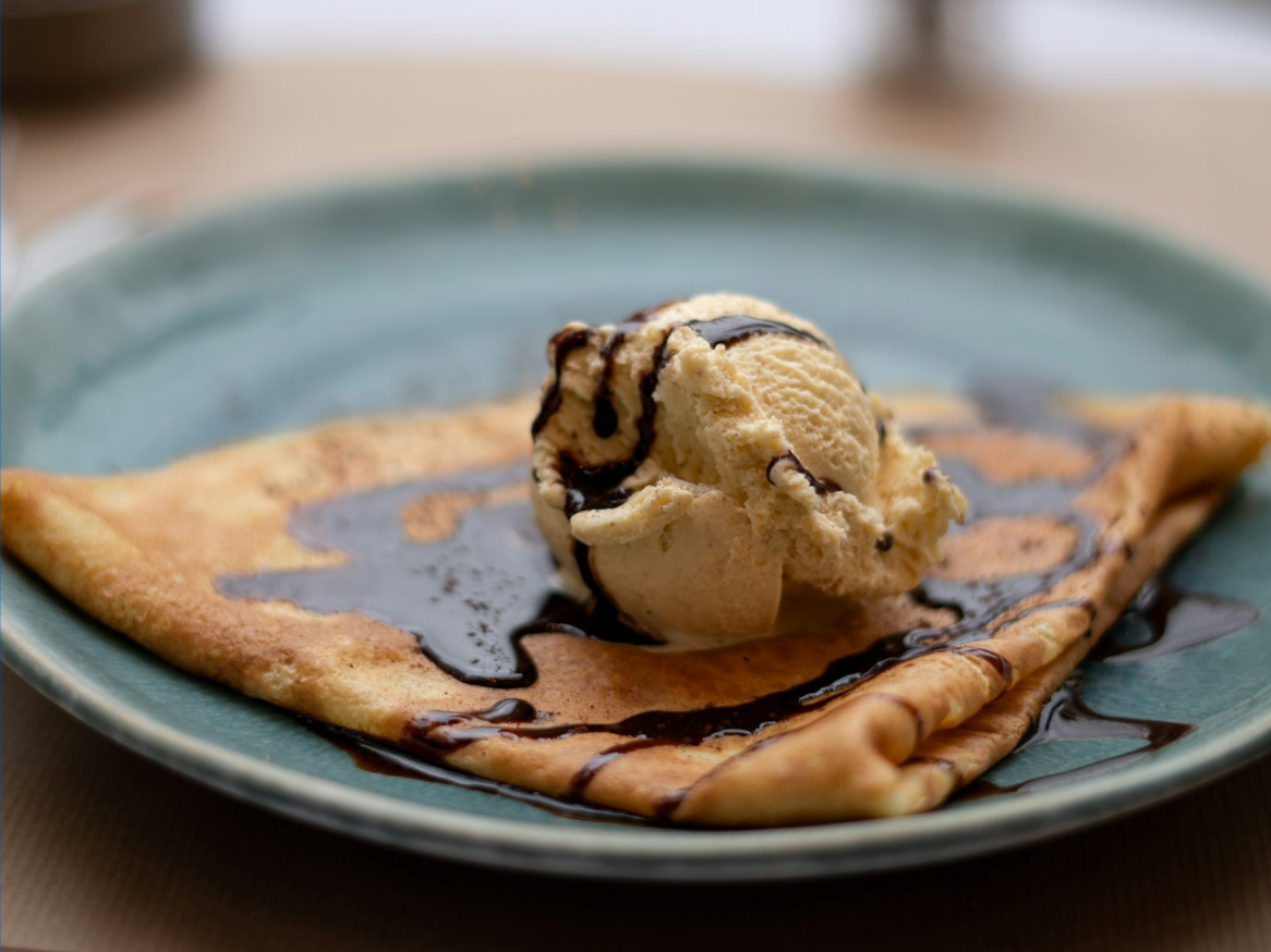 Crepe dobrado com uma bola de gelado de baunilha e calda de chocolate num prato azul.