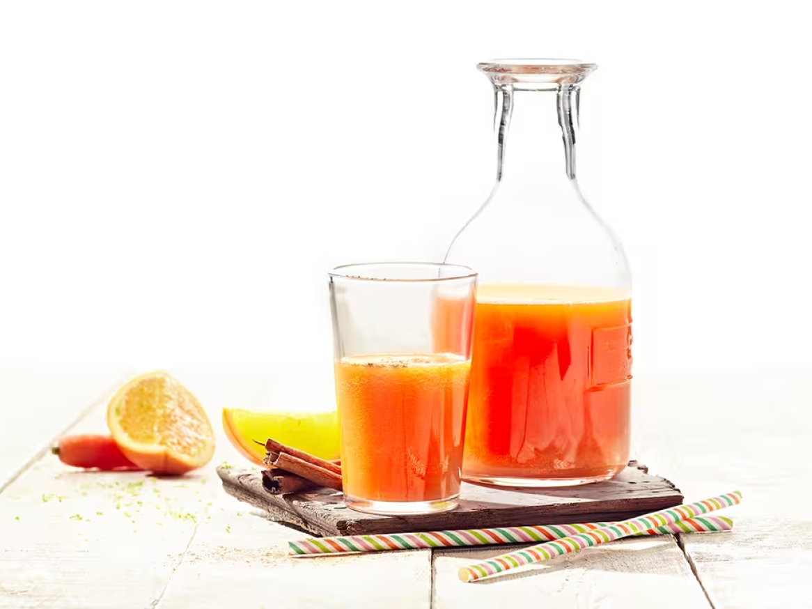 Sumo de cenoura e laranja fresco em copo e jarro, com canela e rodelas de laranja.