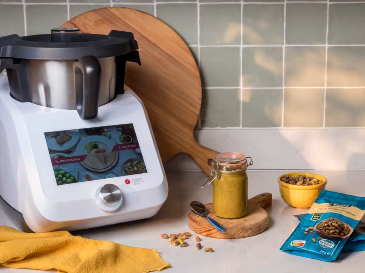 Um robot de cozinha com ecrã tátil, um frasco de pesto de pistácio e pistácios numa bancada de cozinha.
