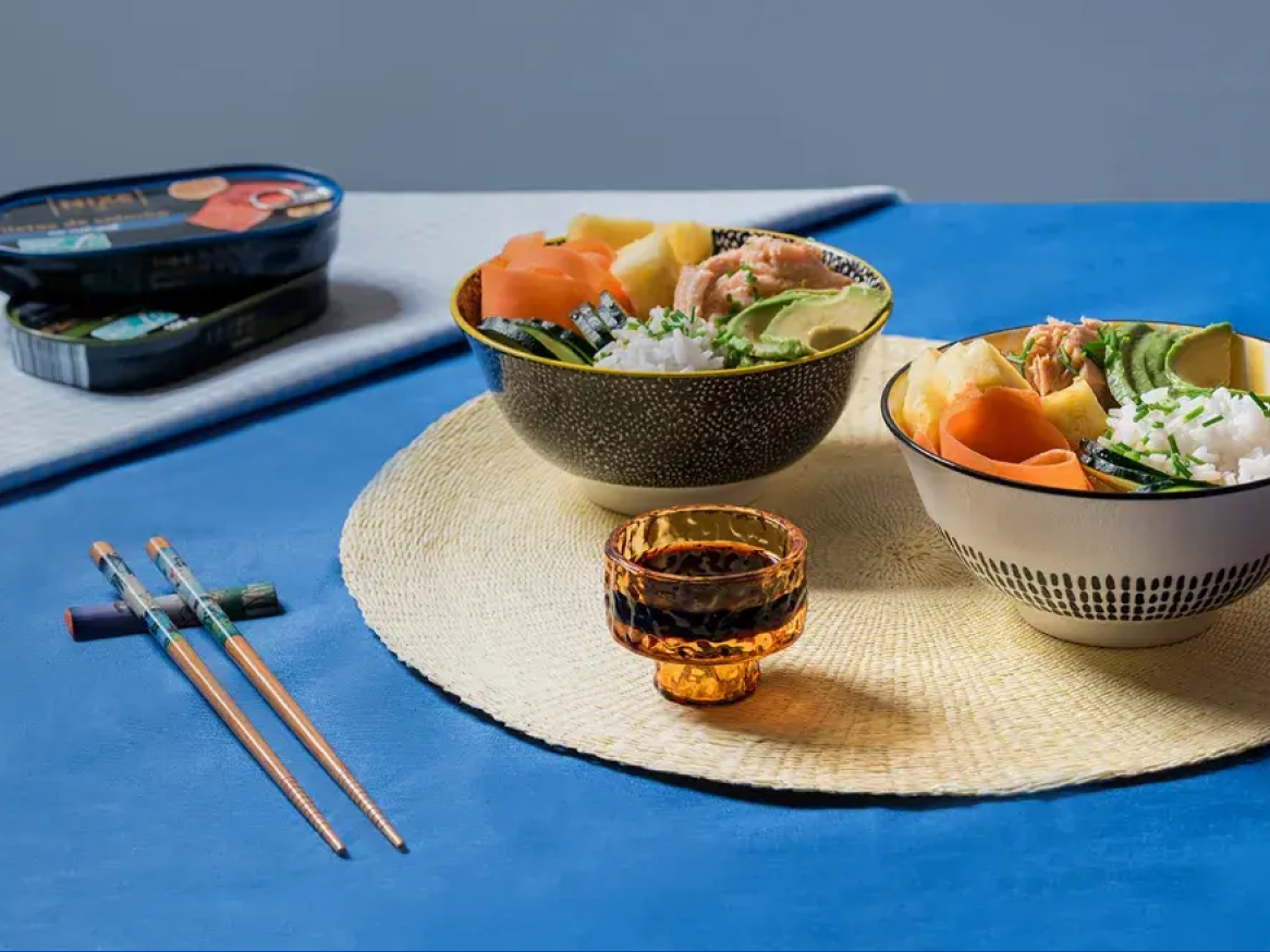 Duas taças de poke com salmão, abacate, arroz e vegetais, com pauzinhos e latas de peixe.