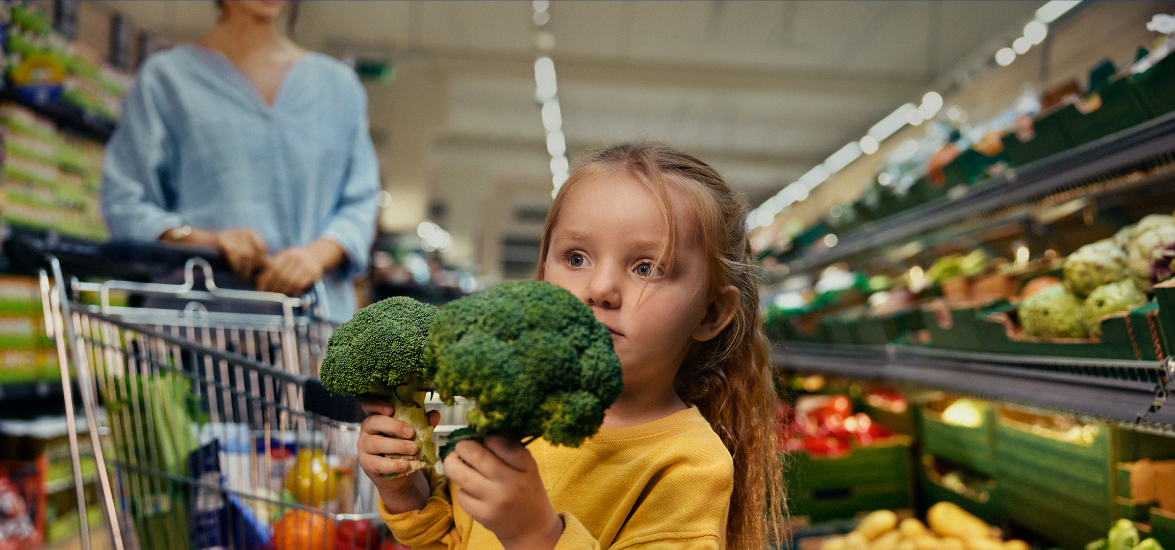 Menina com brócolos no supermercado, com um carrinho de compras e prateleiras ao fundo.
