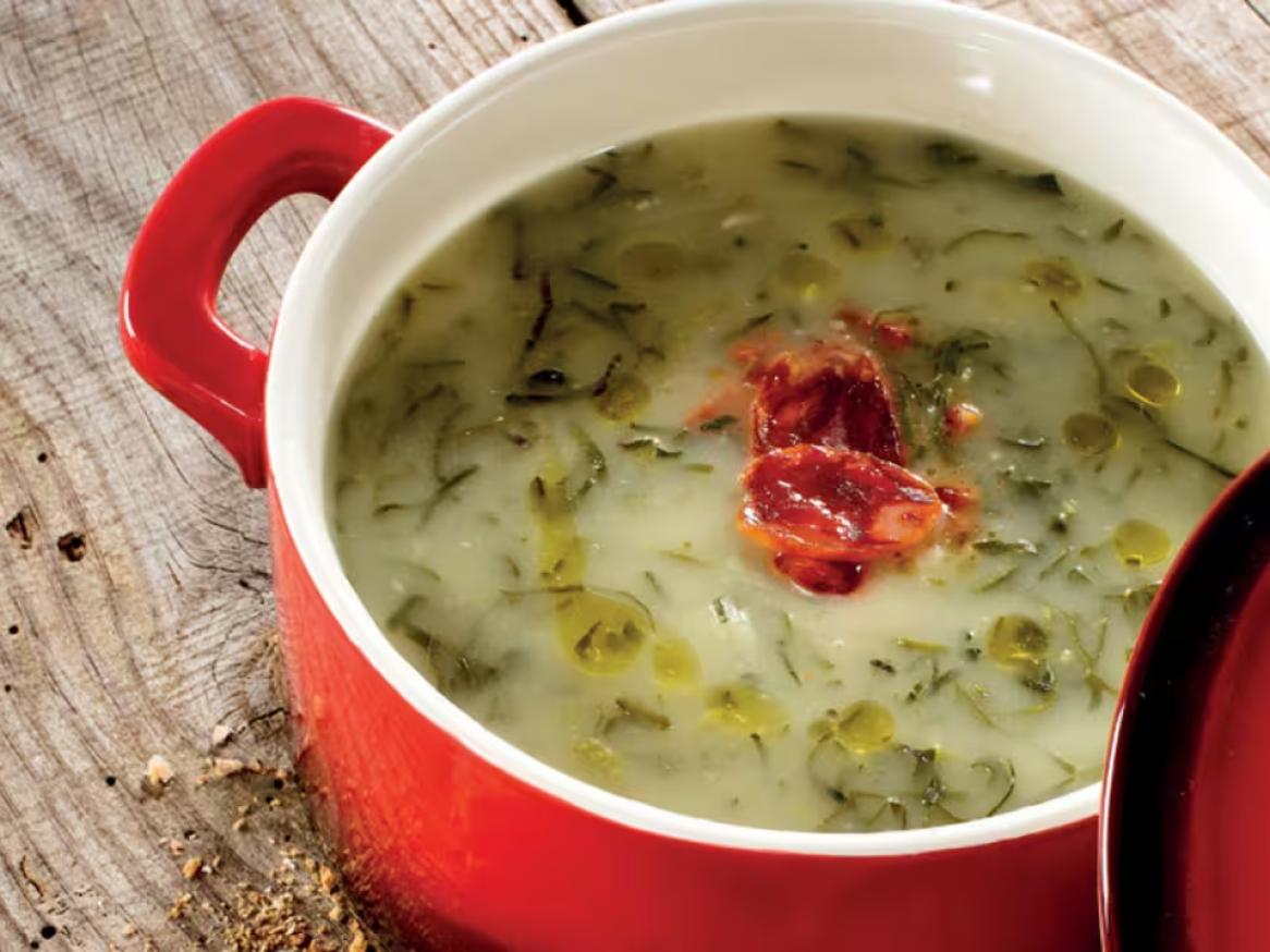 Caldo verde com chouriço numa panela vermelha sobre uma mesa de madeira.