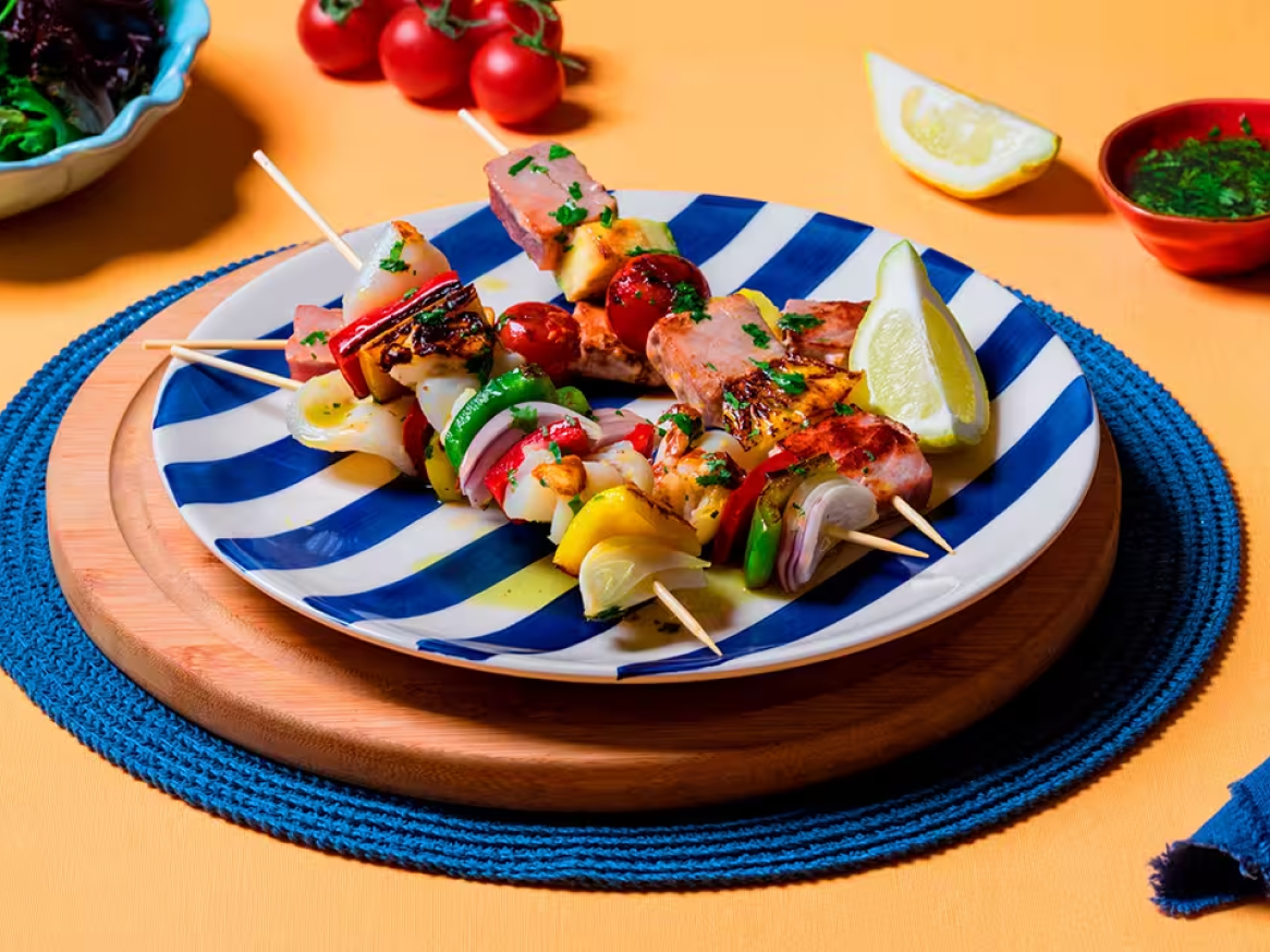 Espetadas de carne e marisco com legumes coloridos, servidas num prato às riscas.