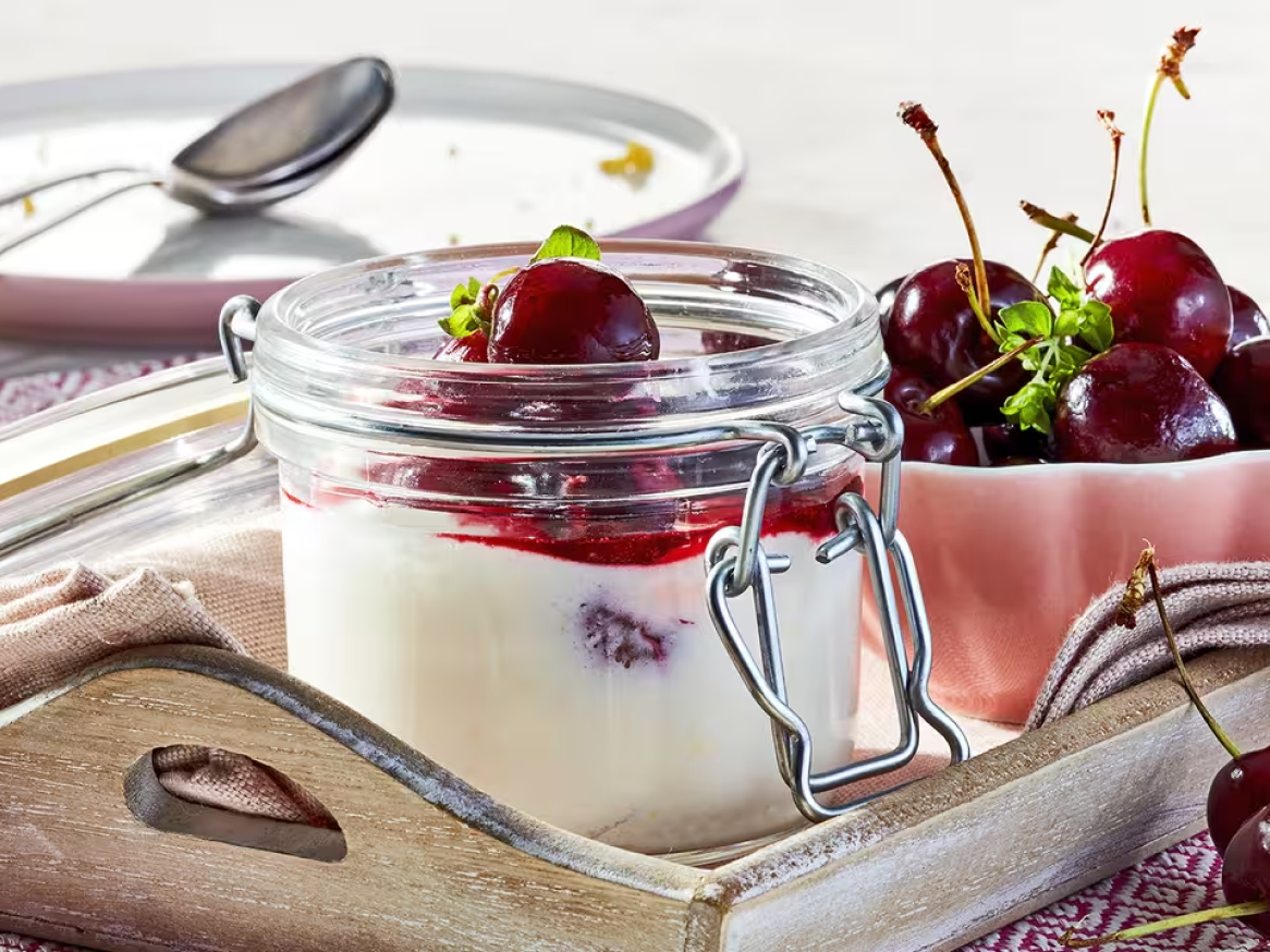 Iogurte com cerejas e molho de cereja num frasco de vidro, com cerejas frescas ao lado.