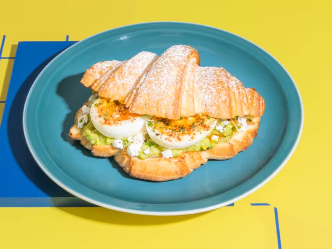 Croissant com abacate, ovo cozido e queijo feta num prato azul, sobre fundo amarelo e azul.