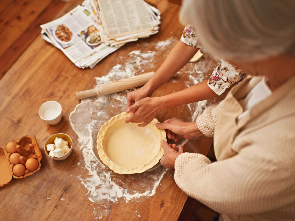 Mãos a preparar uma tarte com massa, ovos, manteiga e farinha numa mesa de madeira.