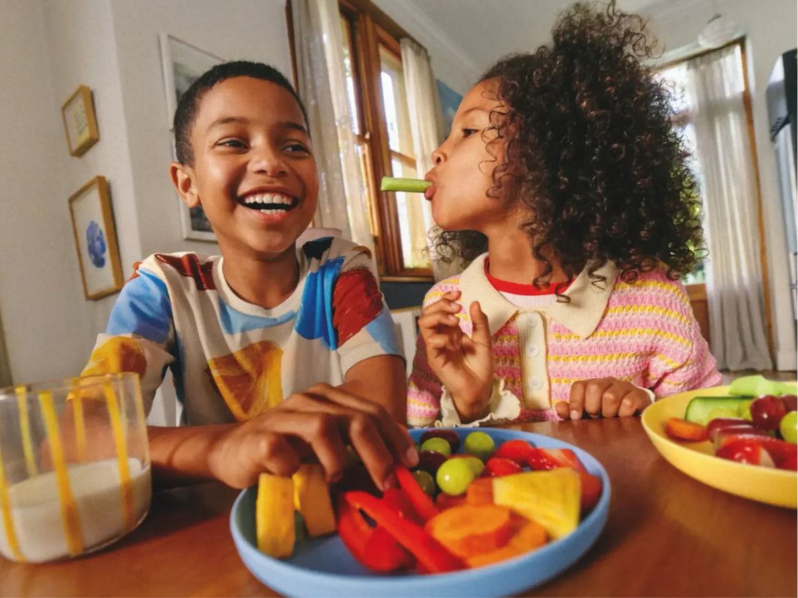 Duas crianças felizes a comer fruta e vegetais frescos à mesa.