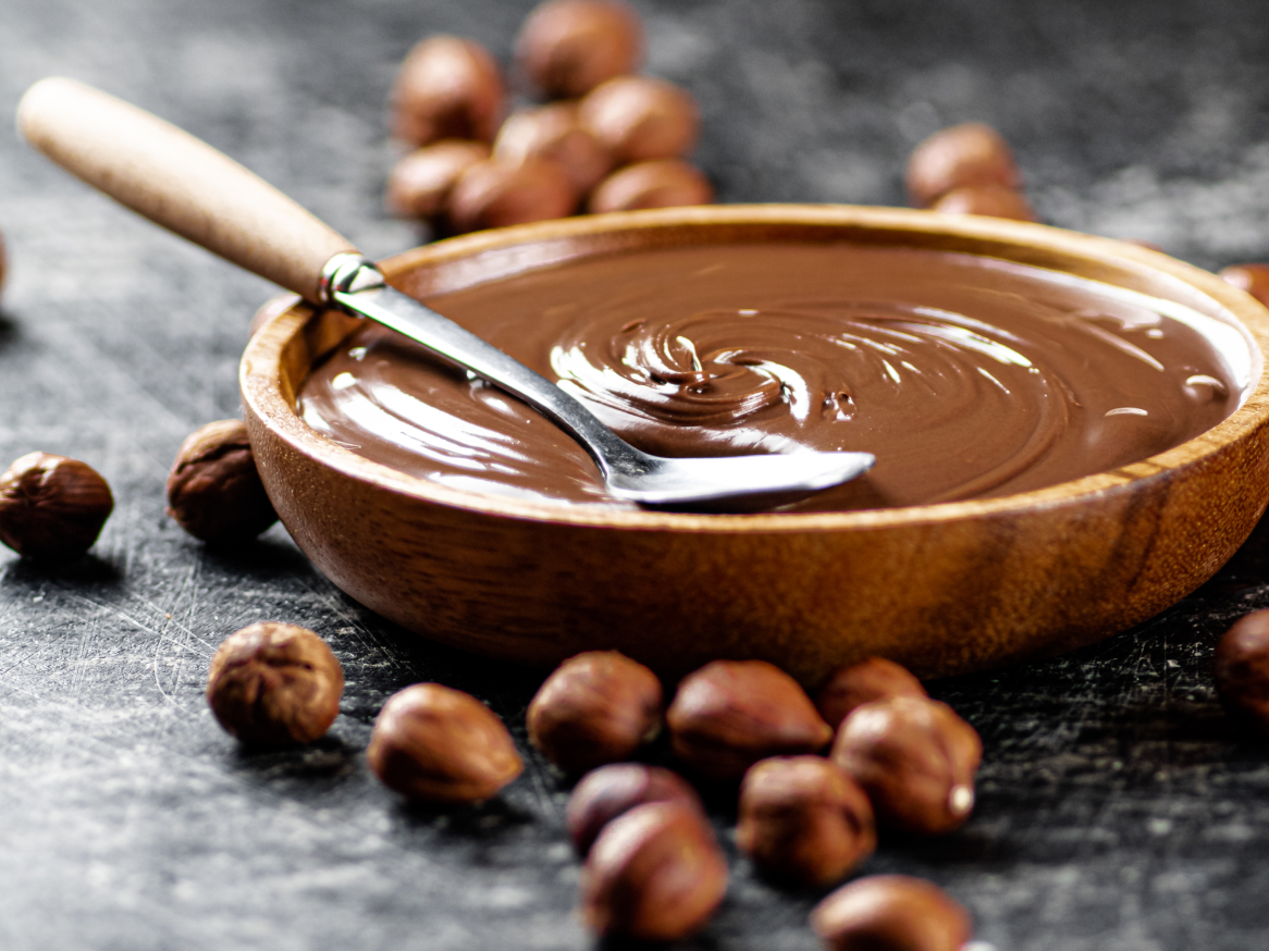 Creme de avelã e chocolate numa tigela de madeira com uma colher, rodeado de avelãs.