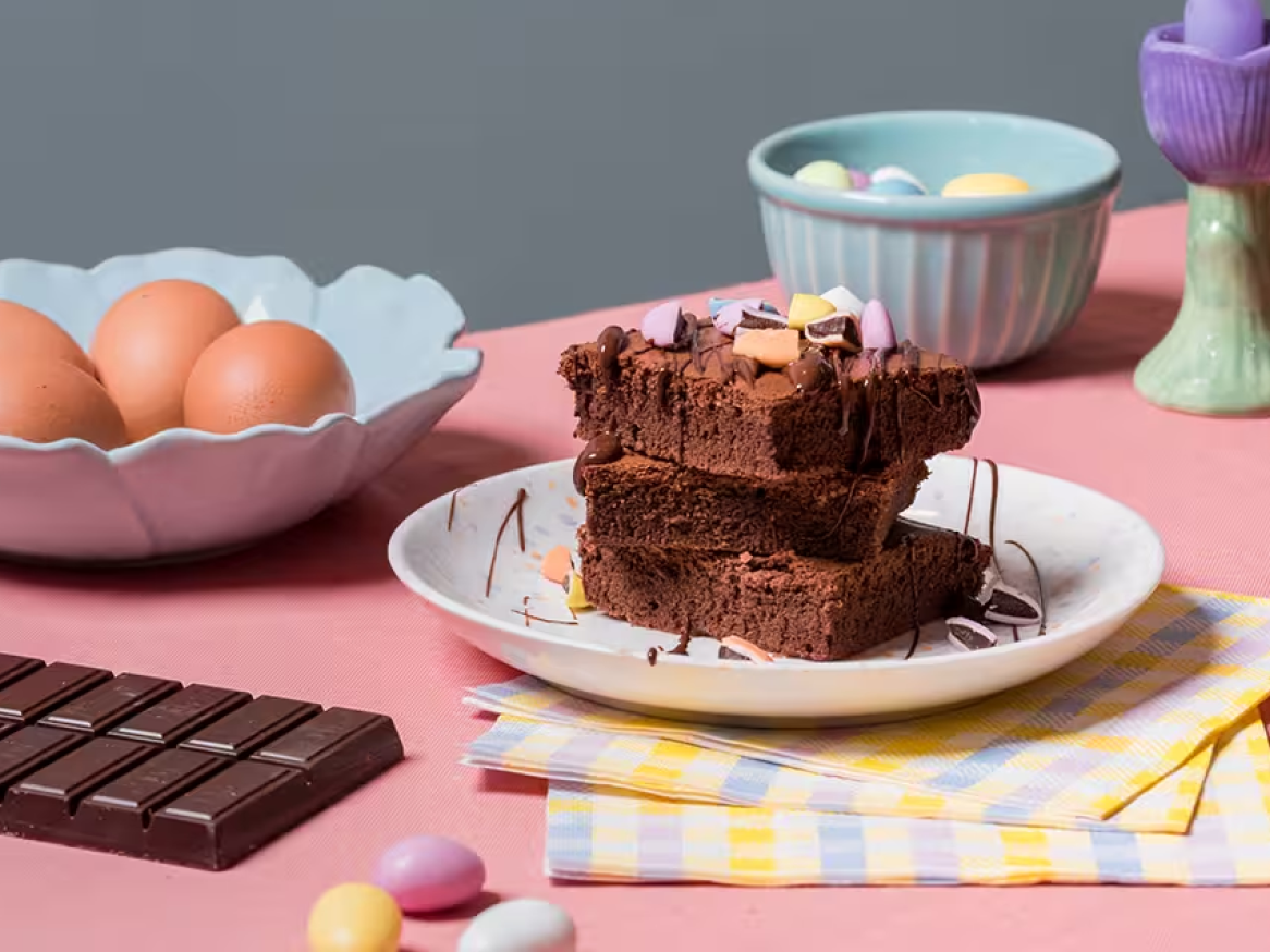 Brownies de chocolate com cobertura, ovos e uma barra de chocolate numa mesa rosa.