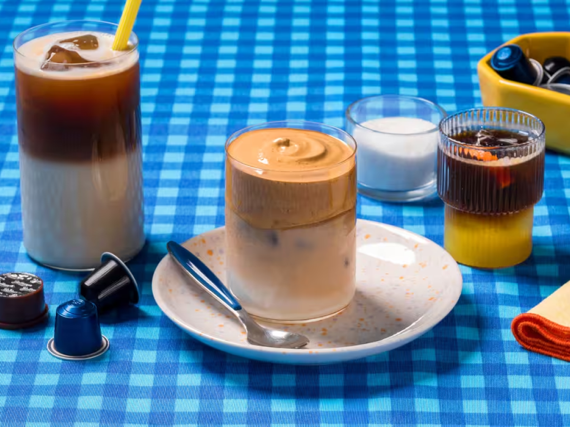 Bebidas de café geladas e cápsulas de café sobre uma toalha de mesa azul xadrez.