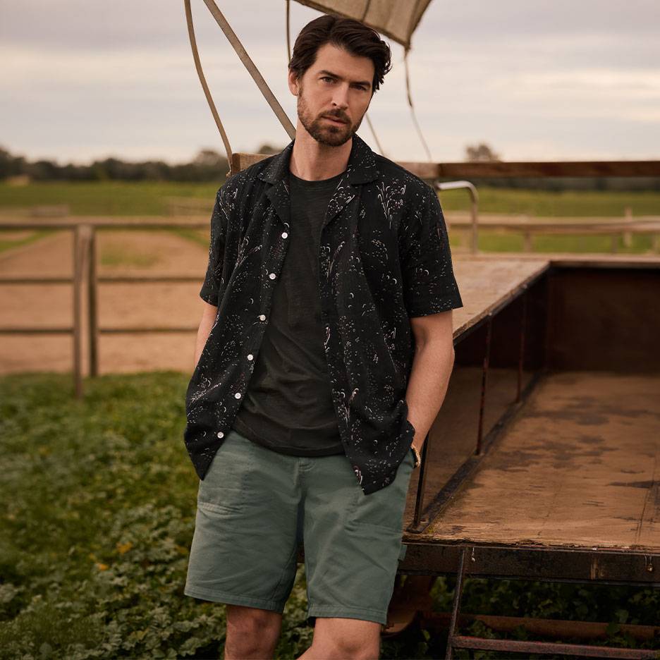 Homem de camisa preta estampada, t-shirt e calções verdes, de pé junto a um reboque.