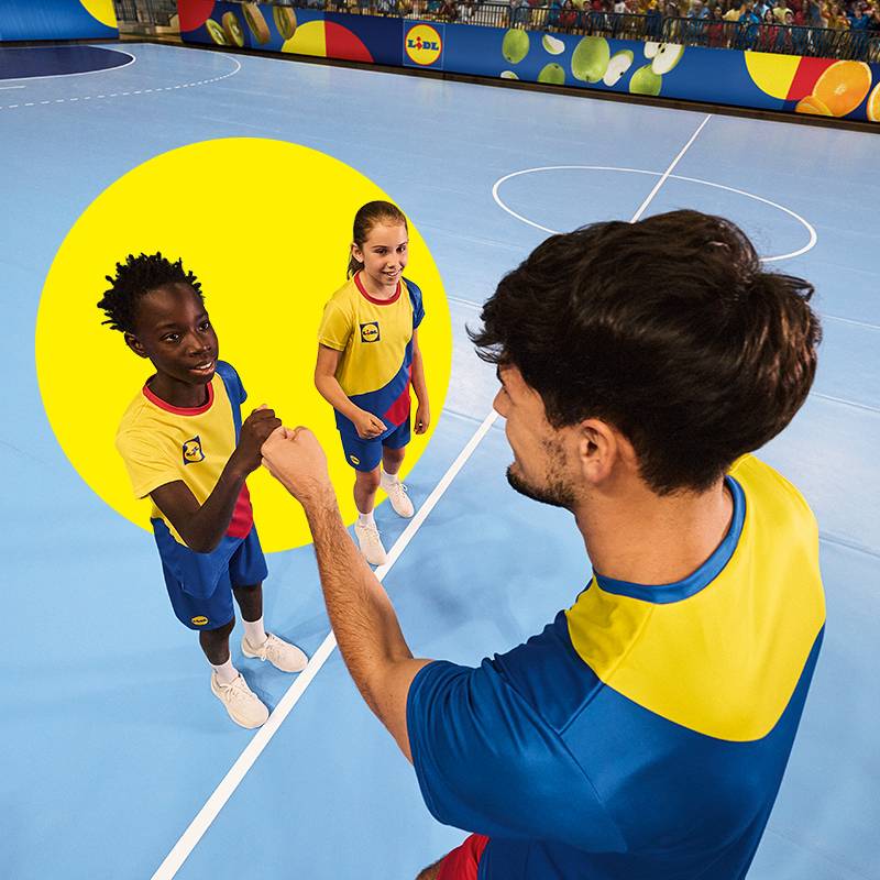 Crianças com roupa desportiva Lidl dão um high-five a um adulto num campo interior.