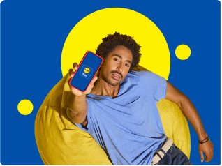 Homem sorridente sentado num pufe amarelo, a mostrar um smartphone com a aplicação de compras.