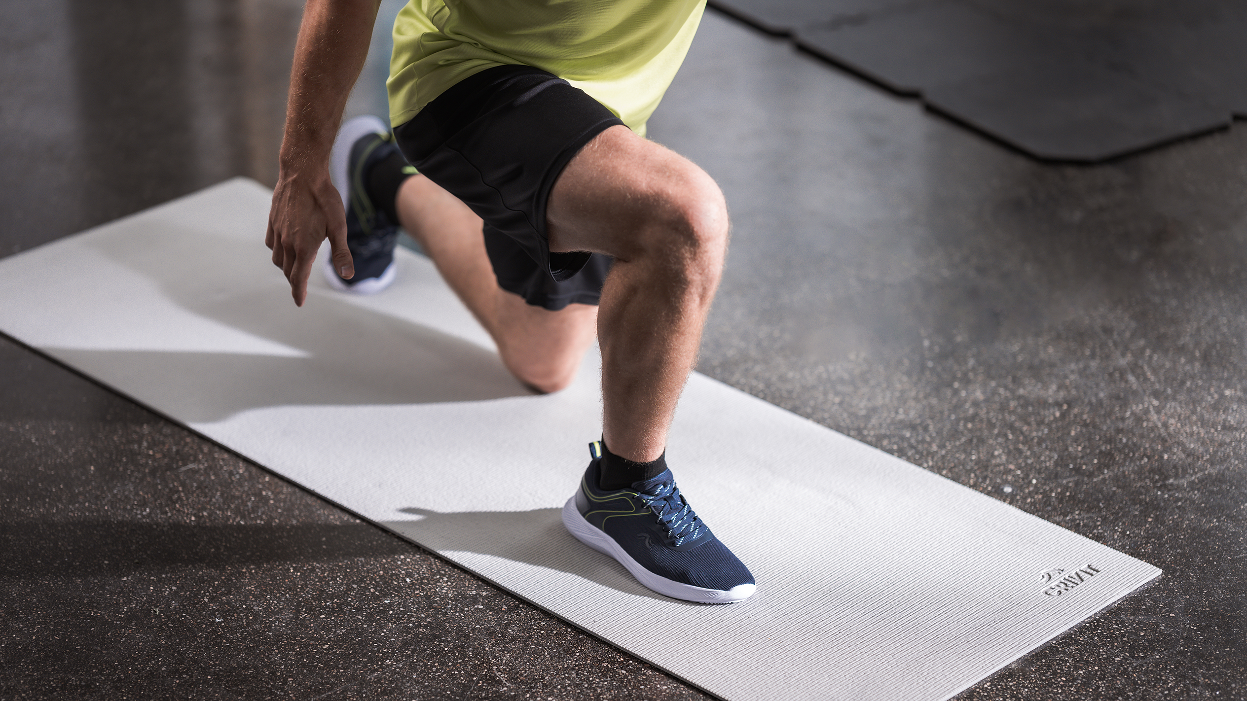 Homem em roupa desportiva a fazer lunges num tapete de exercício cinzento claro.