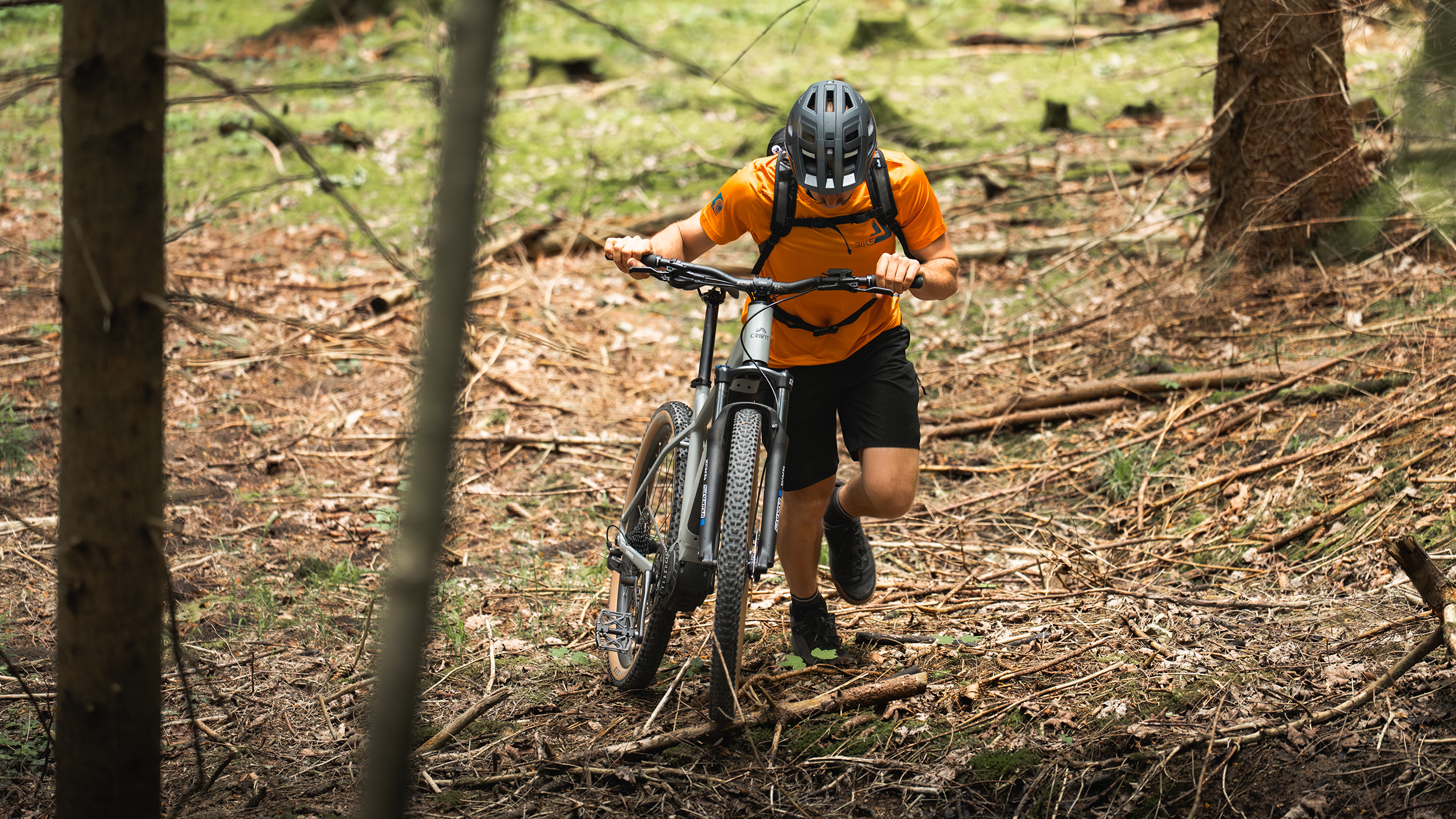 Um ciclista com uma camisa laranja empurra uma bicicleta elétrica de montanha por uma floresta.