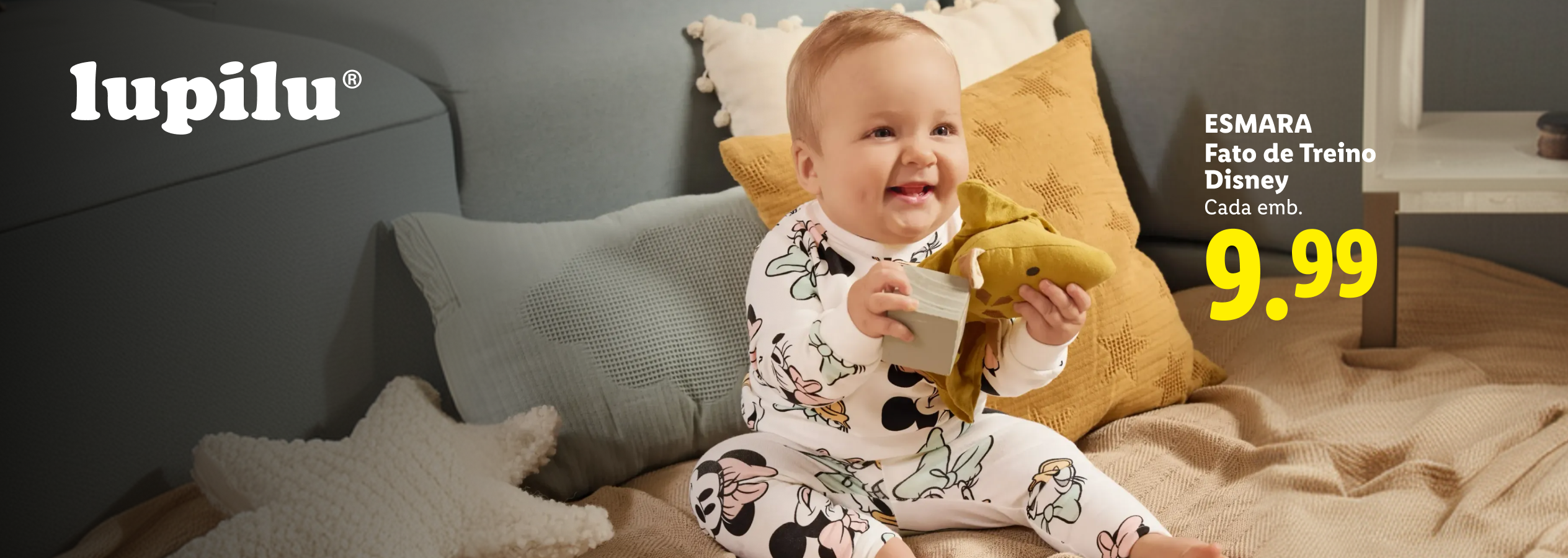 Bebé sorridente em pijama com padrão de personagens de desenhos animados, segurando um brinquedo e um bloco, com o preço de 9,99€.