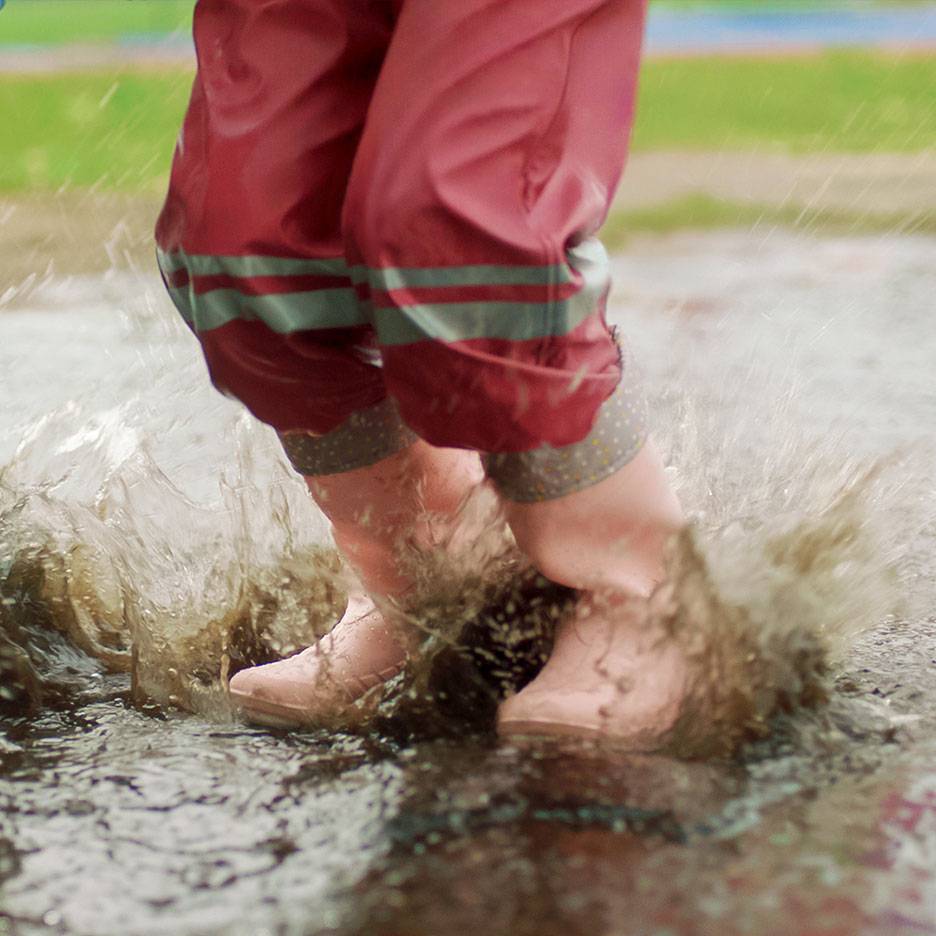 Criança com calças impermeáveis vermelhas e botas a saltar numa poça.