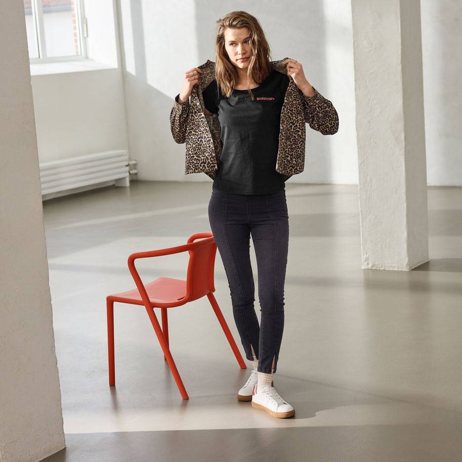 Mulher com t-shirt preta com texto, jeans e casaco com estampa de leopardo, em pé ao lado de uma cadeira vermelha.