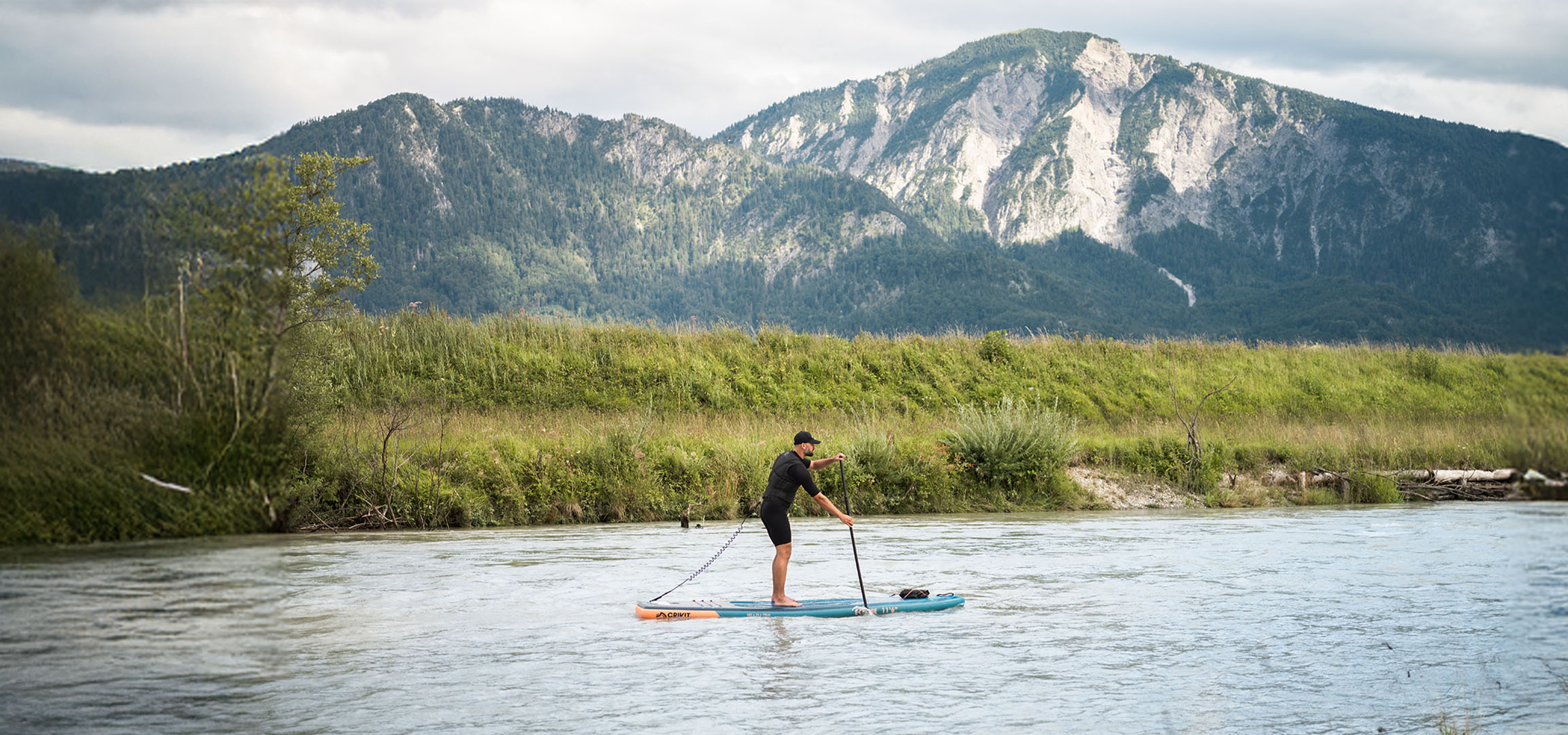 Homem a fazer paddleboard num rio com montanhas e vegetação exuberante ao fundo.
