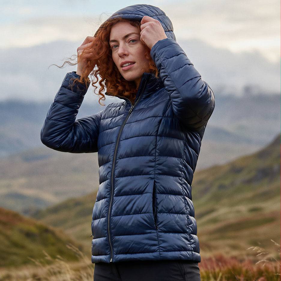 Mulher com cabelo ruivo num casaco acolchoado azul escuro com capuz numa paisagem montanhosa.