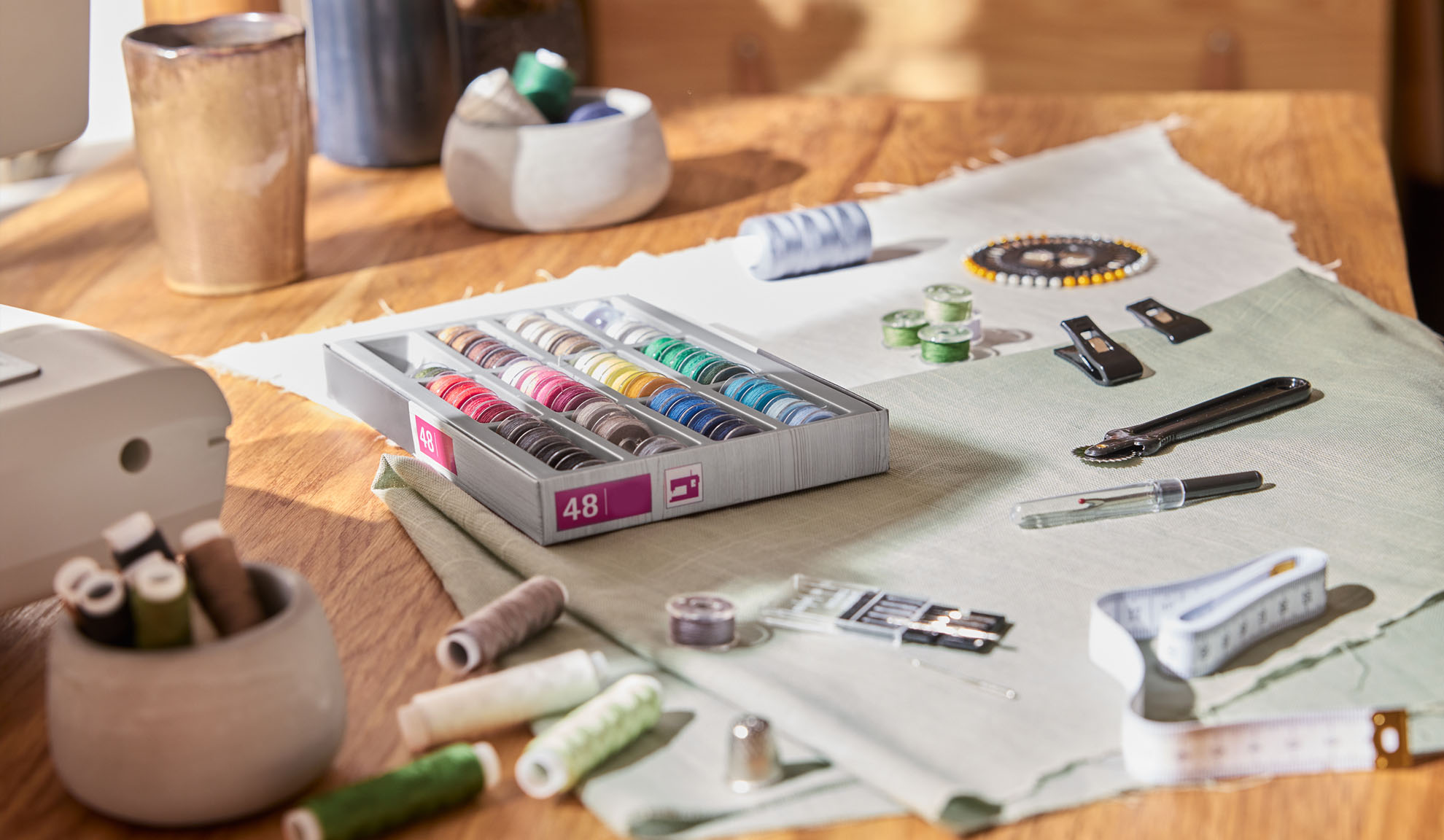 Kit de costura com caixa de linhas coloridas, máquina de costura e várias ferramentas numa mesa de madeira.