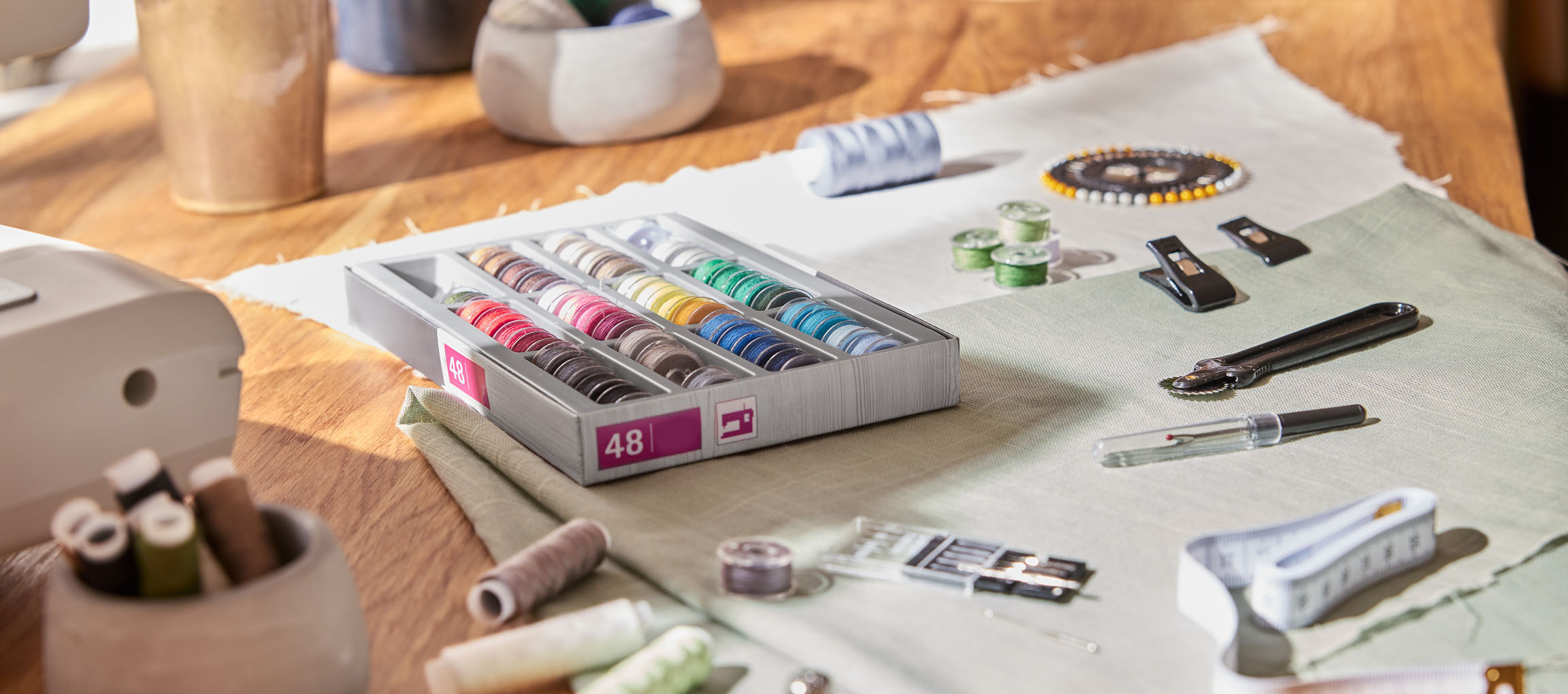 Artigos de costura numa mesa de madeira, incluindo um organizador de linhas com 48 bobinas, tecido e várias ferramentas.