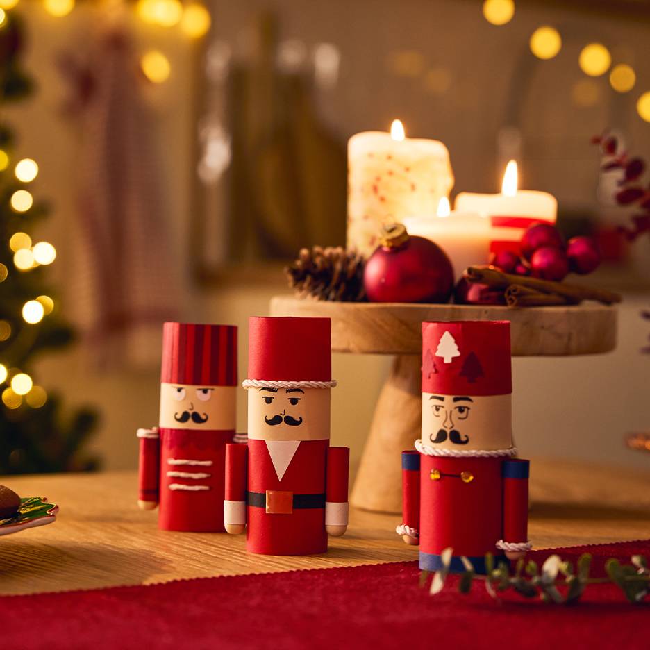Três quebra-nozes de papel festivos numa mesa com decorações de Natal e velas acesas.
