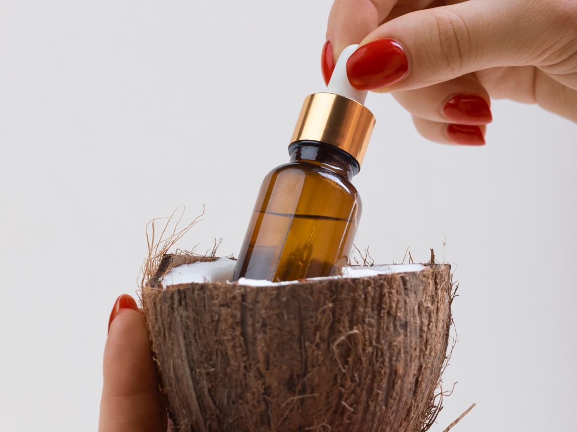 Óleo de coco em frasco conta-gotas dentro de um coco partido, segurado por uma mão.