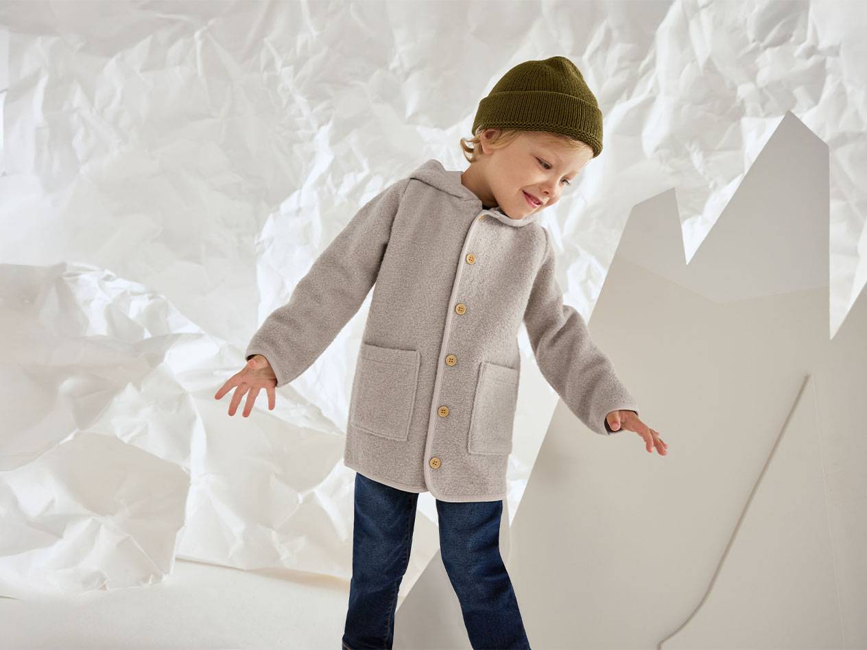 Menino com casaco de lã cinzento e gorro verde, sorrindo contra um fundo de papel amassado.