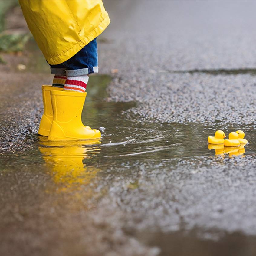 Criança de galochas amarelas e capa de chuva numa poça com patinhos de borracha.