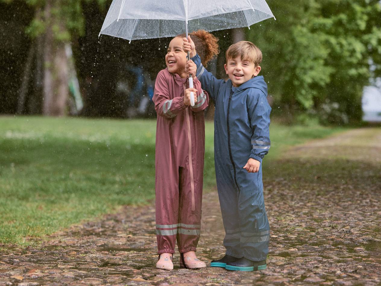 Duas crianças felizes em fatos de chuva, debaixo de um guarda-chuva transparente.