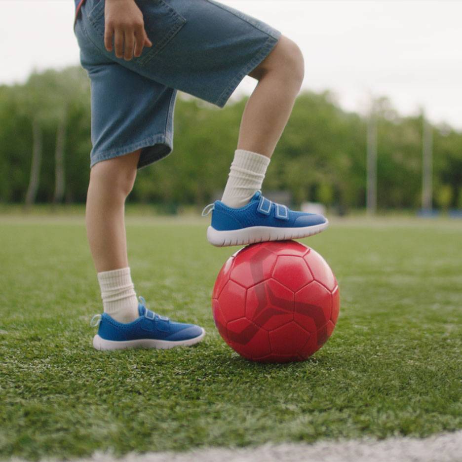 Criança de calções e sapatilhas azuis, com um pé numa bola de futebol vermelha.