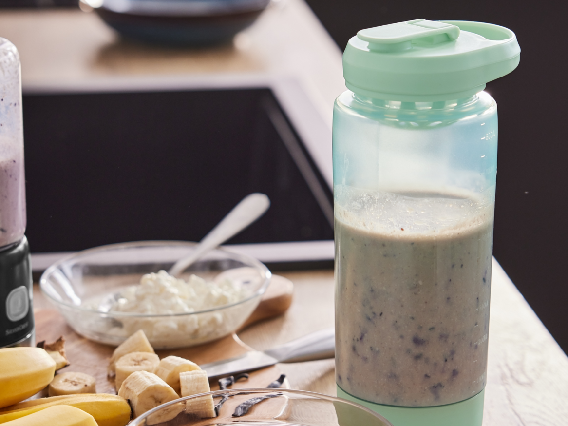 Um liquidificador portátil com um smoothie, bananas e queijo cottage em uma bancada.