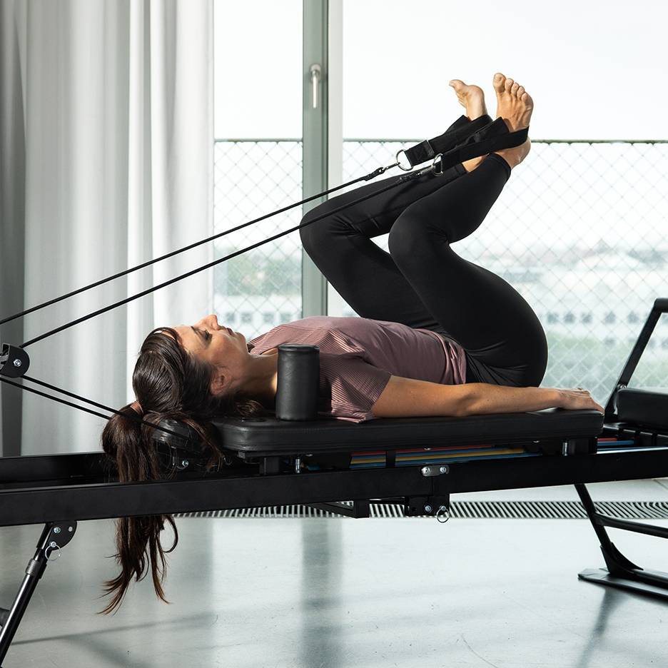 Mulher a exercitar as pernas num reformer de Pilates com bandas de resistência.