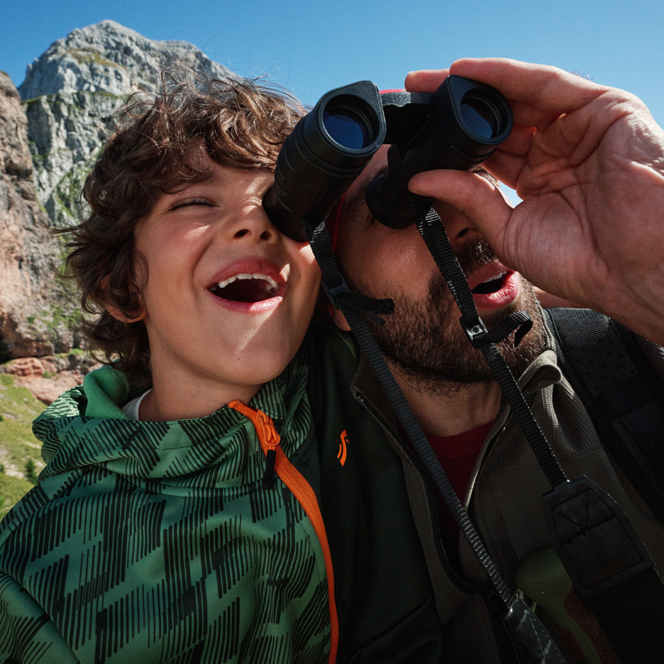 Pai e filho explorando montanhas com binóculos, usando casacos de exterior.