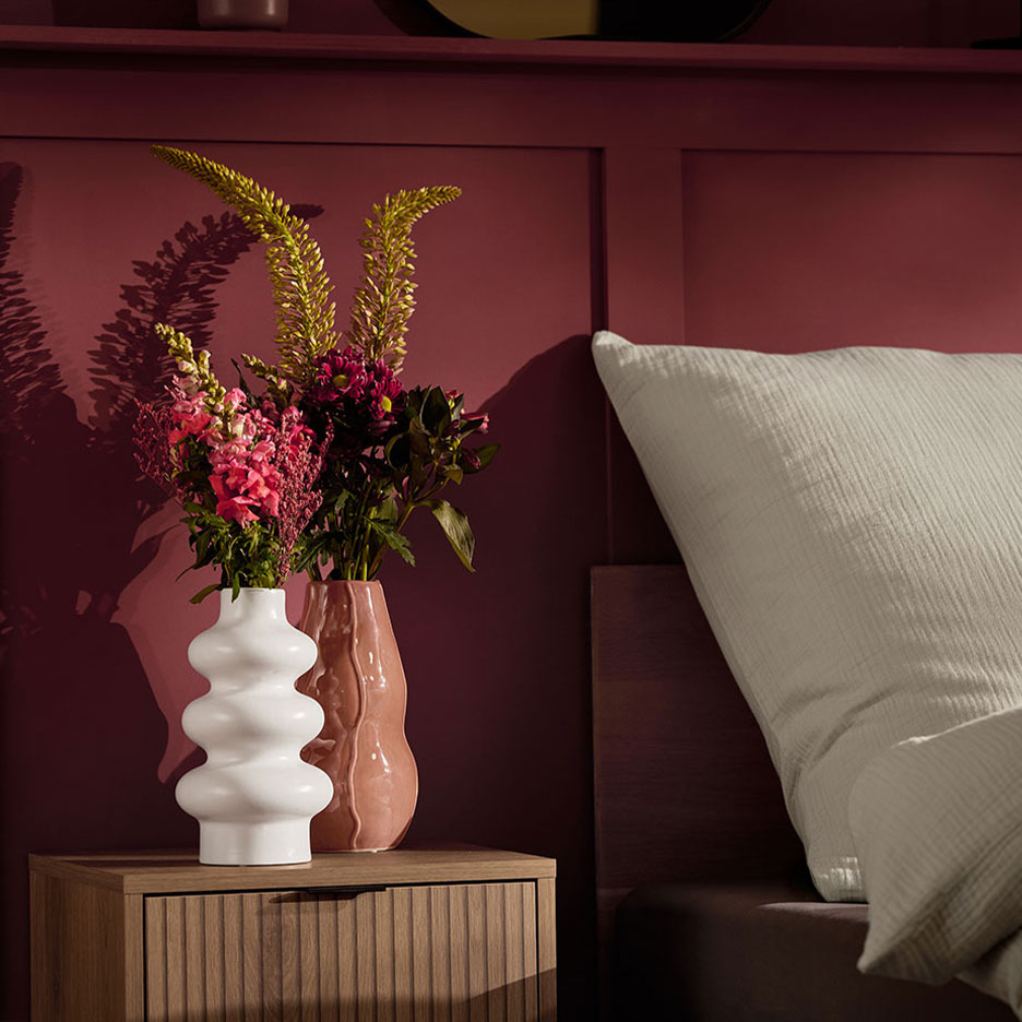 Duas jarras com flores numa mesa de cabeceira ao lado de uma cama com almofada branca.