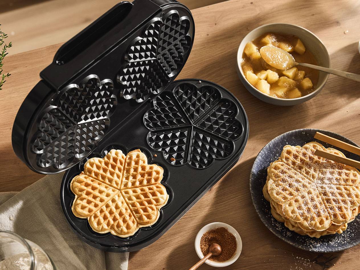 Uma máquina de waffles aberta com um waffle em forma de coração, ao lado de waffles cozidos e compota de maçã.