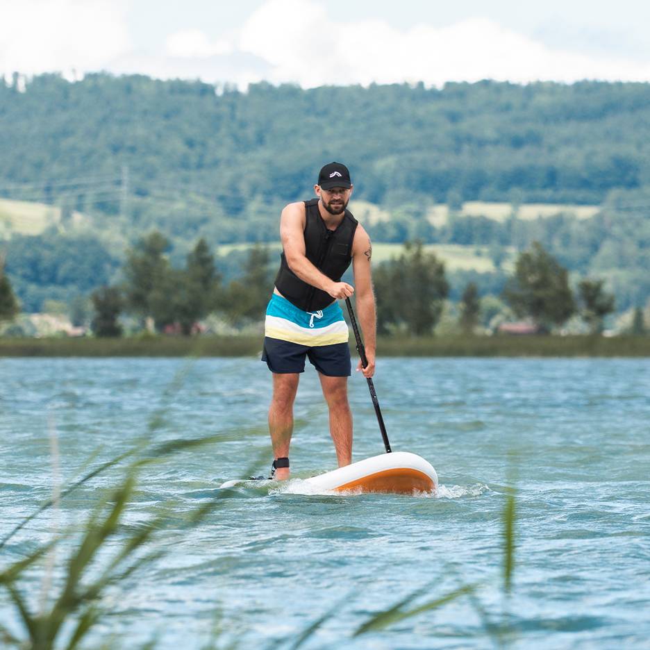 Homem a fazer stand-up paddle num lago, com colete salva-vidas e calções de banho.