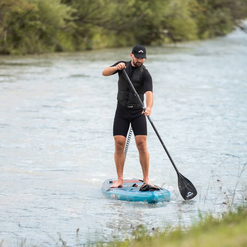 Homem a praticar paddleboard num rio, vestindo fato de mergulho preto, colete salva-vidas e boné.