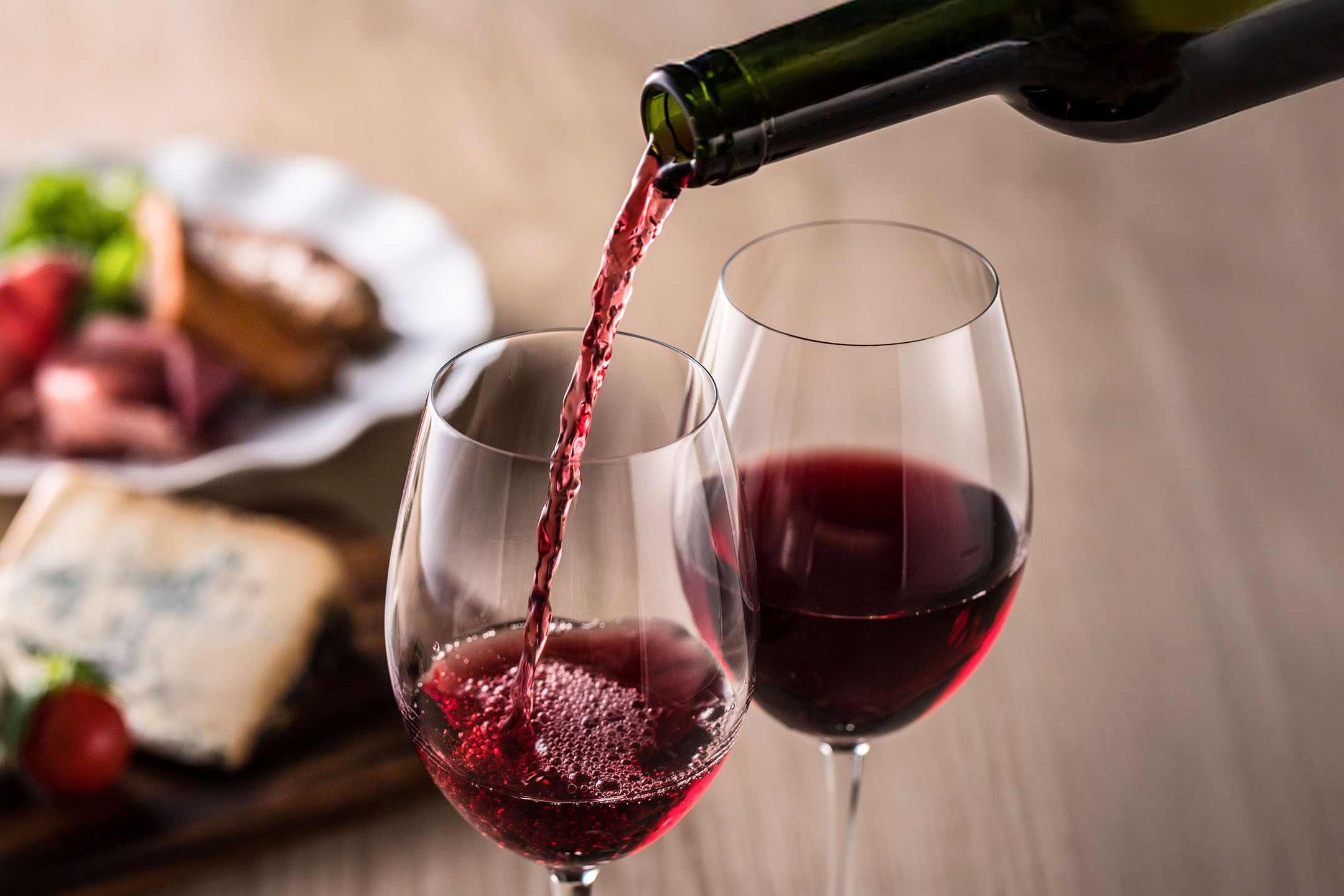 Vinho tinto a ser servido num copo, com aperitivos ao fundo.