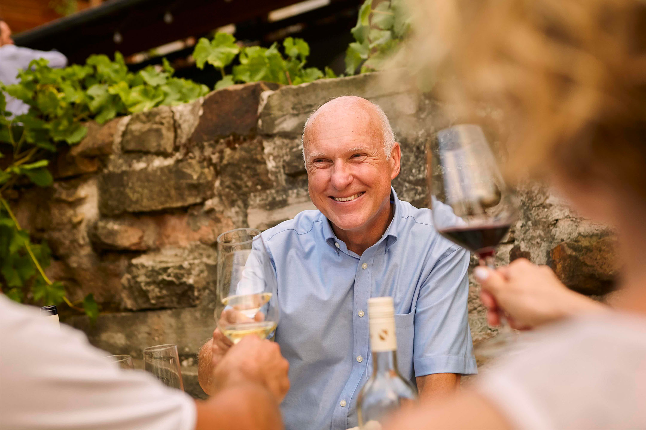 Homem sorridente a brindar com vinho branco, rodeado de amigos e uma parede de pedra.