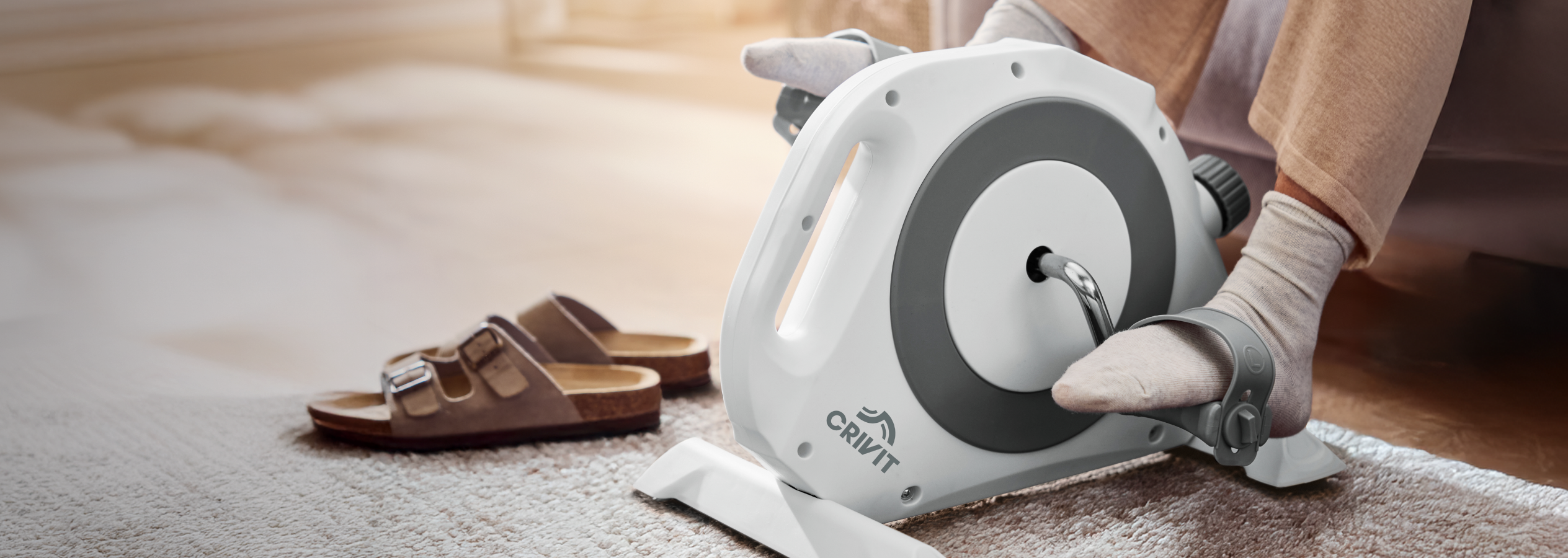 Pés a pedalar um mini-bicicleta ergométrica Crivit em carpete, com sandálias ao lado.