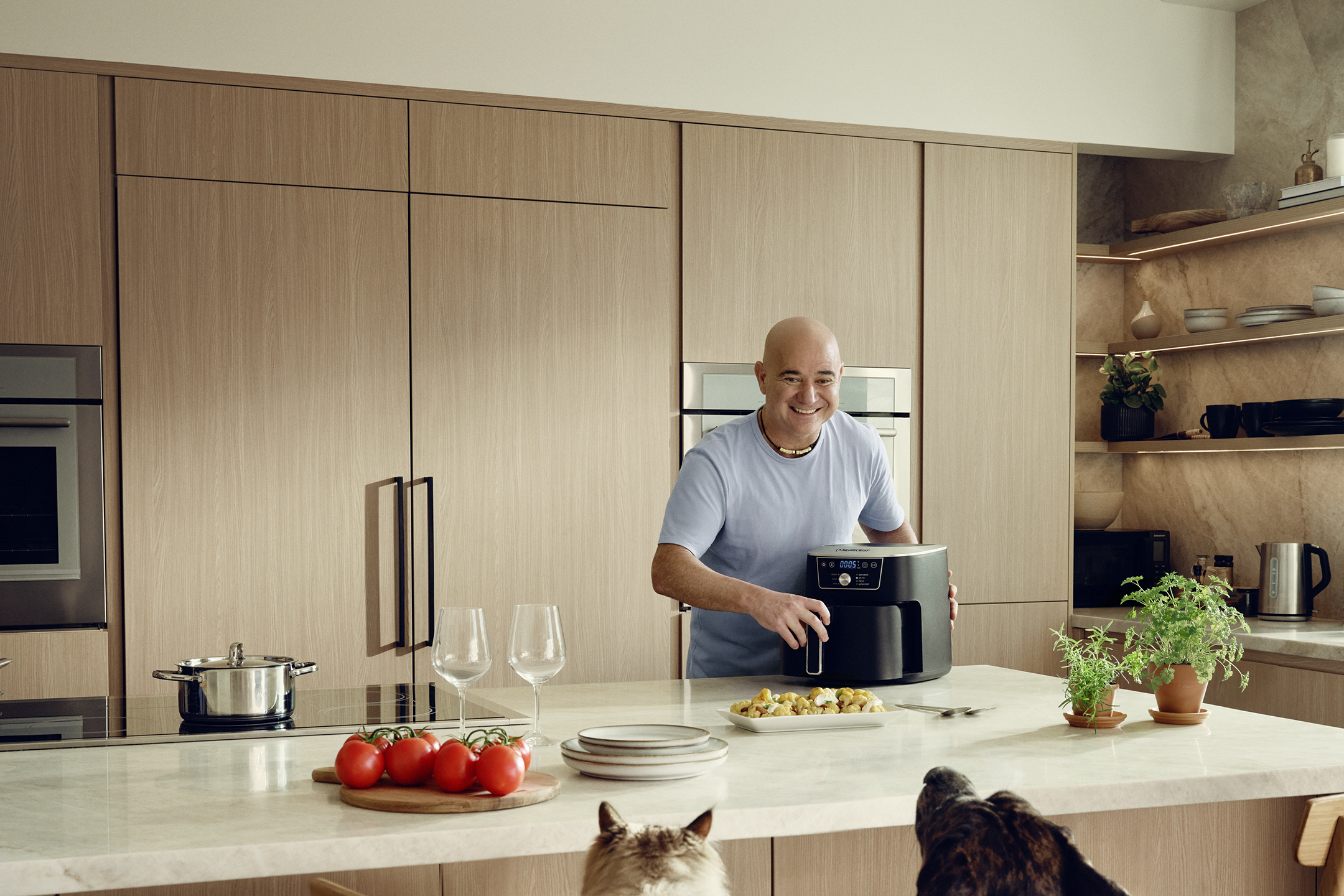 Homem na cozinha com fritadeira de ar, preparando uma refeição enquanto cães observam.